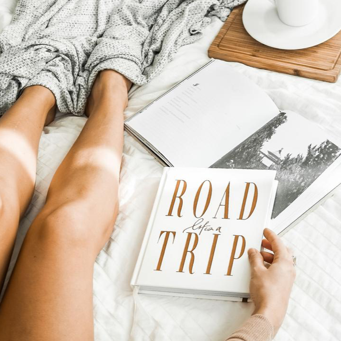 A woman's legs are on a bed with the book "Life's A ROADTRIP - Luxe Edition" from the AXEL & ASH range.
