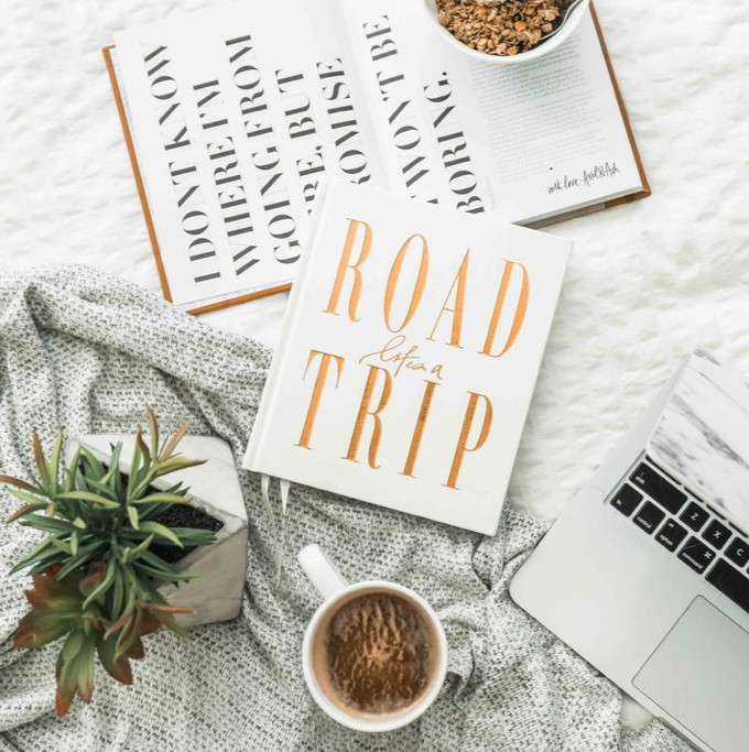 An AXEL & ASH Life's A ROADTRIP - Luxe Edition book, accompanied by a laptop and coffee, featuring the words "road trip" on its cover.