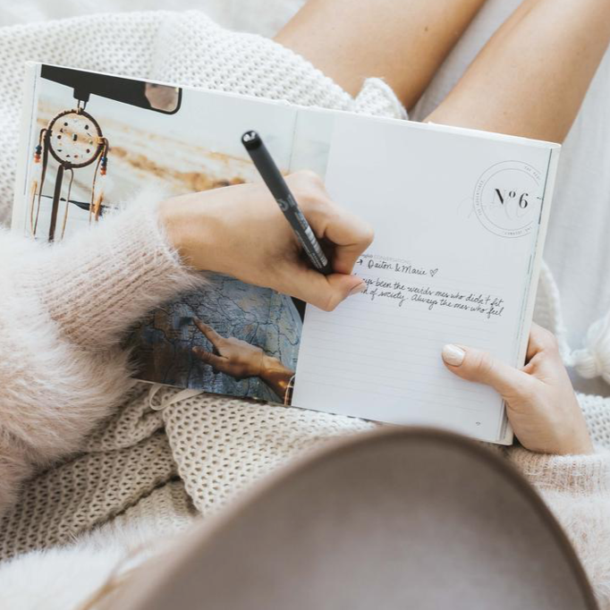 A woman jotting down notes in a Life's A ROADTRIP - Luxe Edition notebook by AXEL & ASH on a cozy bed.