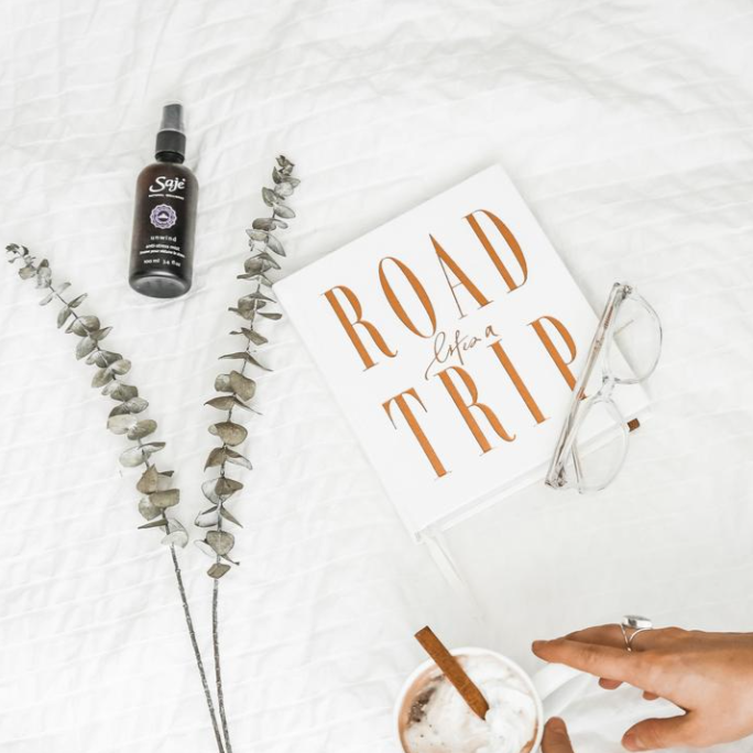 A hand with a cup of coffee and a bottle of Life's A ROADTRIP - Luxe Edition essential oil from the AXEL & ASH range on a bed.
