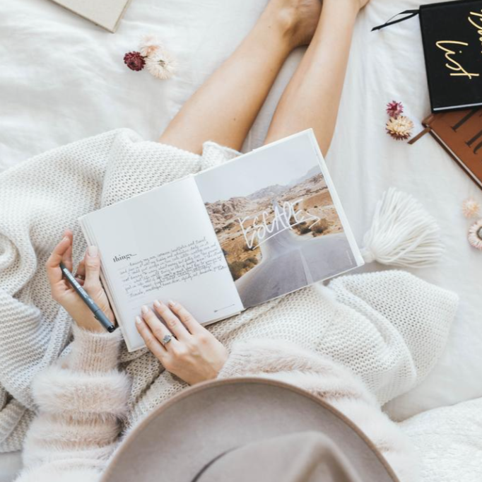 A woman lounging on a bed with a Life's A ROADTRIP - Luxe Edition book from AXEL & ASH.