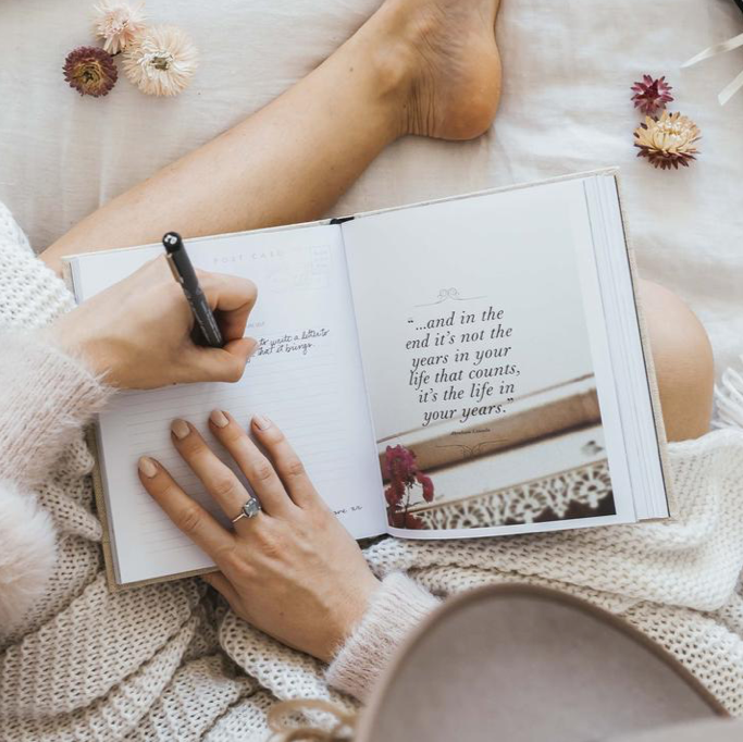 A woman with a vintage feel, writing in a WANDERLUST travel journal on a bed from AXEL & ASH.