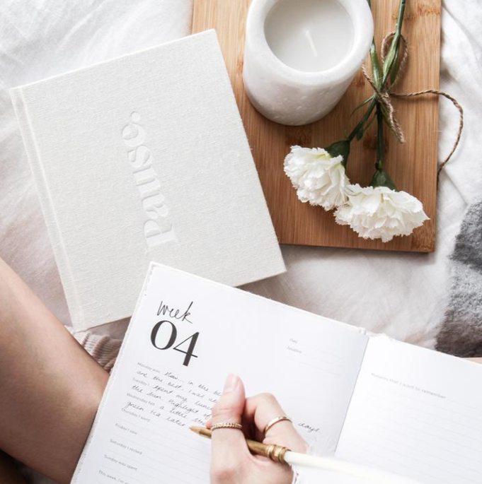 Hand writing in Press Pause, open at a week 04 page, beside a candle and white flowers