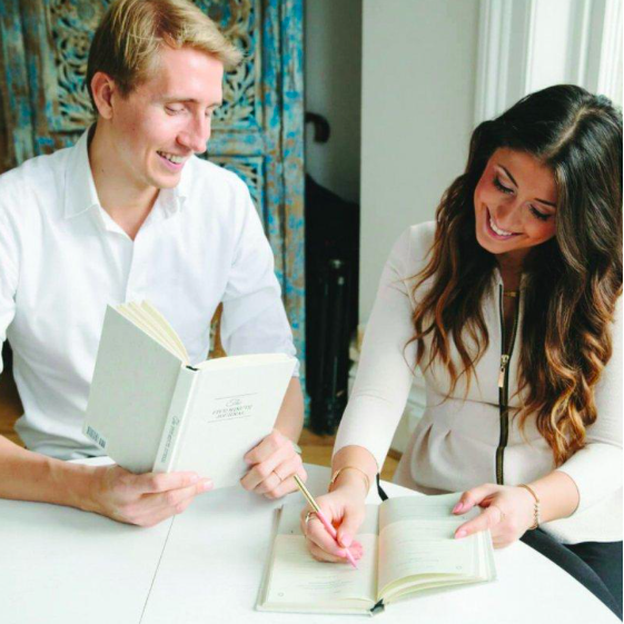 Man and woman sharing a laugh while filling in their journals at a white table