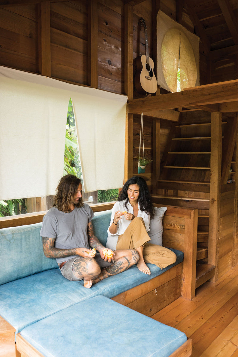 An artistic couple sitting on a surf shack couch in a cabin, surrounded by Surf Shacks: An Eclectic Compilation Of Creative Surfers Homes From Coast To Coast and Overseas books.