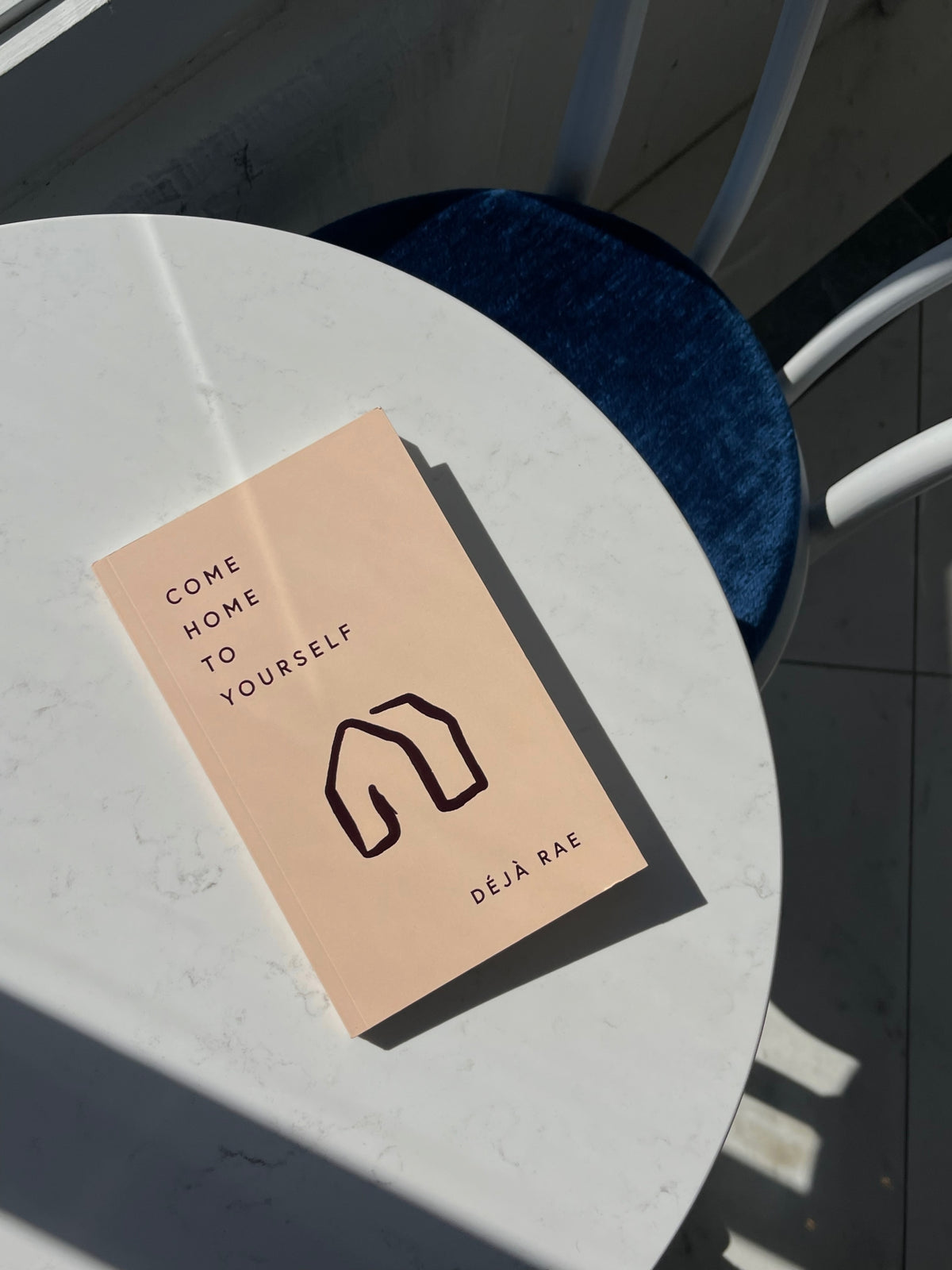 A Come Home to Yourself book by Déjà Rae is sitting on a table next to a window. (Brand: Thought Catalog)