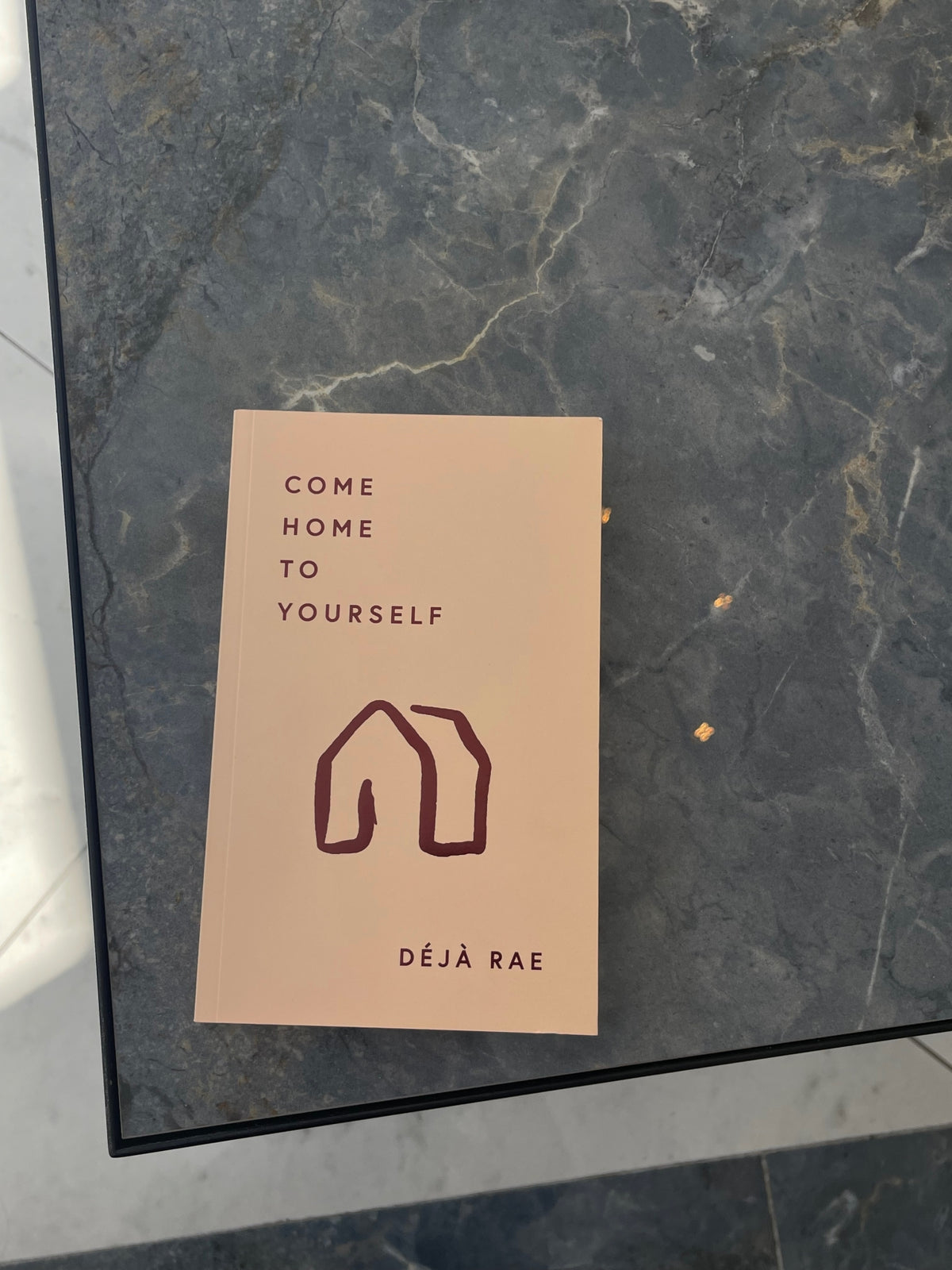 A book titled "Come Home to Yourself | Déjà Rae" by Thought Catalog that explores solitude and relationships, displayed on a sleek marble table.