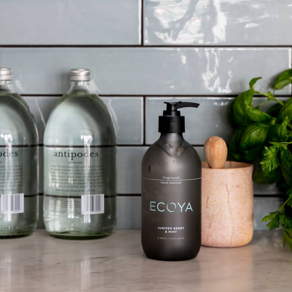 A bottle of Ecoya fragranced hand sanitiser in the Kitchen scent, with its sleek design, sits on a counter next to a bottle of water, making it a perfect Scandinavian gift option.