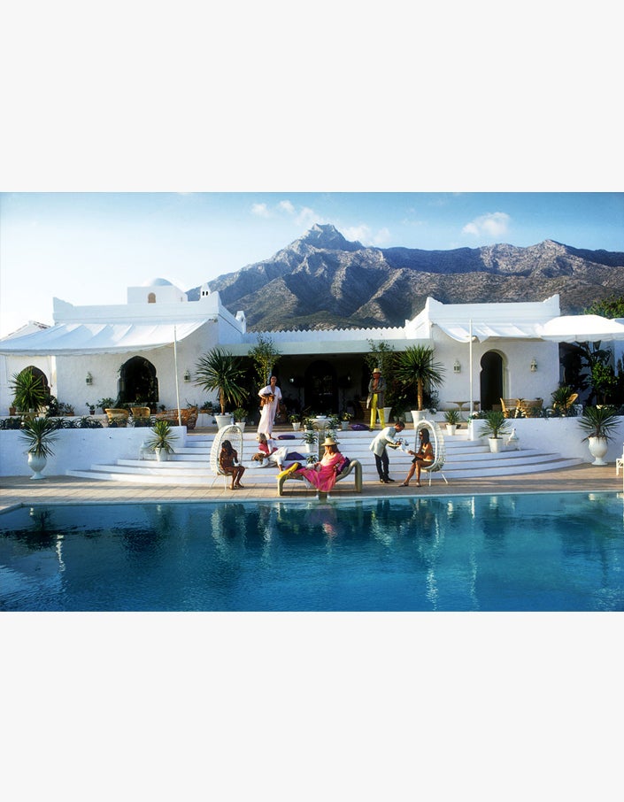 A group of people sitting around a Poolside with Slim Aarons book with mountains in the background.