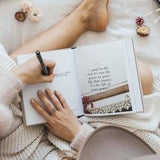 A woman is sitting on a bed and writing a travel journal with Swept Away by WANDERLUST, an AXEL & ASH product, giving it a vintage feel.