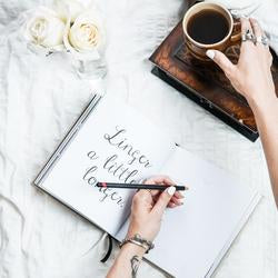 A vintage-inspired woman writing in a travel journal, Swept Away by WANDERLUST, on a cozy bed with a cup of coffee from AXEL & ASH.