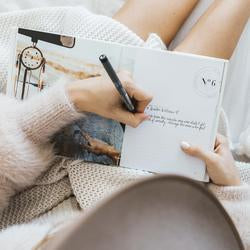 A woman is sitting on a bed, engrossed in the process of writing in the Life's A ROADTRIP - Luxe Edition notebook from the AXEL & ASH range.