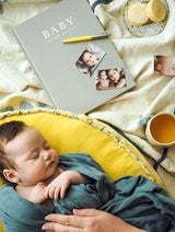 A baby is laying on a yellow blanket next to a Write To Me Baby Journal - Birth To Five Years - Grey / Oatmeal / Pink / Blue.