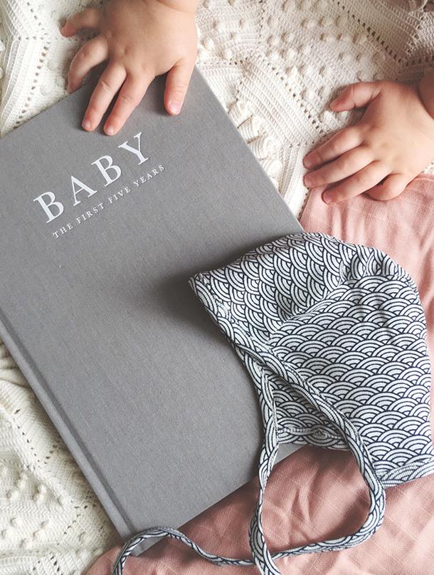 Grey linen Write To Me Baby journal beside a baby's hands and a knitted bonnet