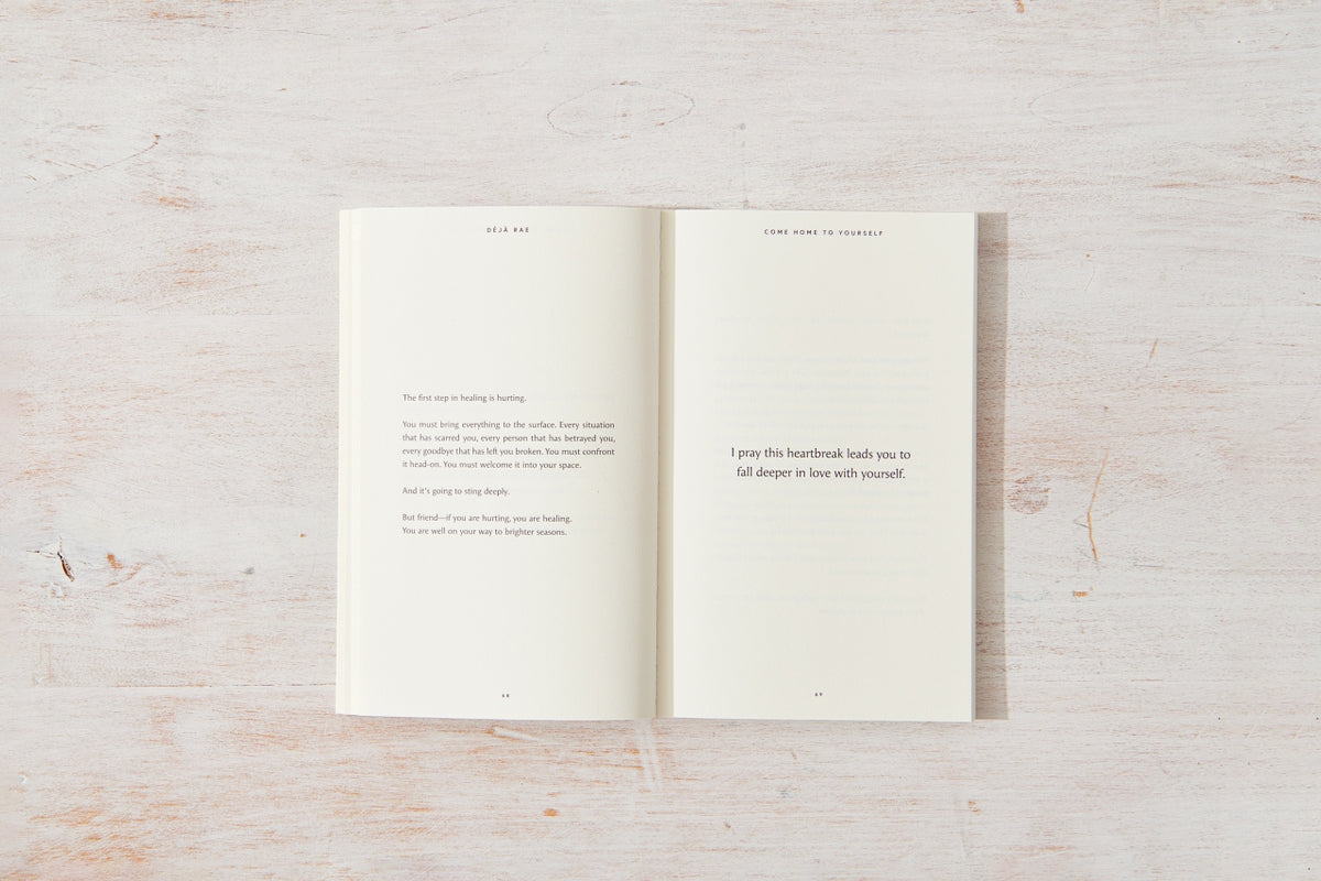A "Come Home to Yourself | Déjà Rae" design book with an open page on a wooden surface by Thought Catalog.
