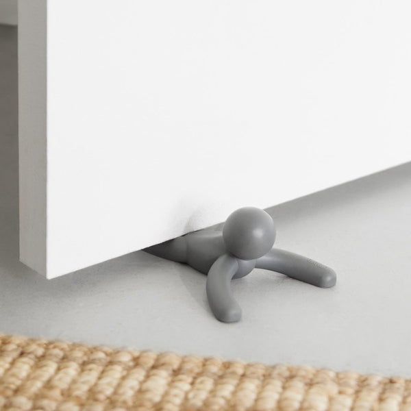 A small grey Buddy Doorstop Charcoal man sitting on the floor of a door. (Brand Name: Umbra)