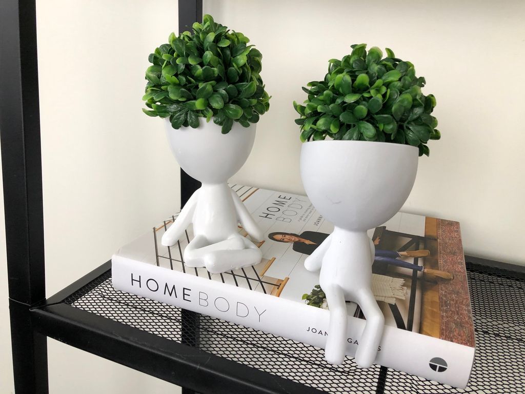 Two white egg head planters holding faux boxwood, perched on a book on a black wire shelf