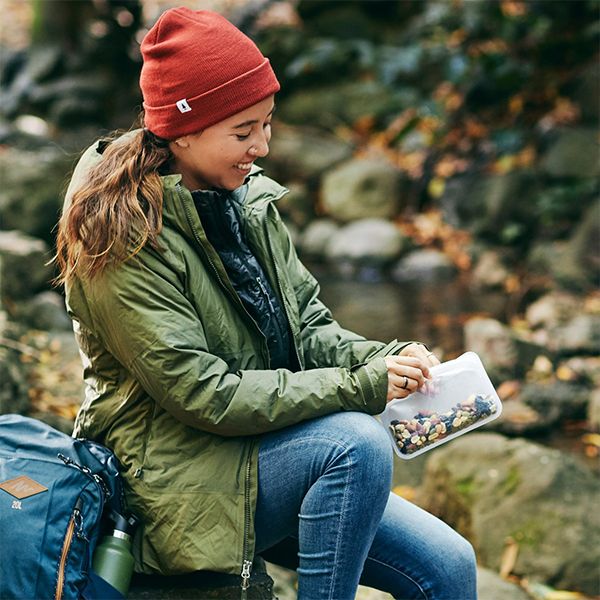 Woman in a red beanie sitting on river rocks, opening a Stasher snack bag of trail mix