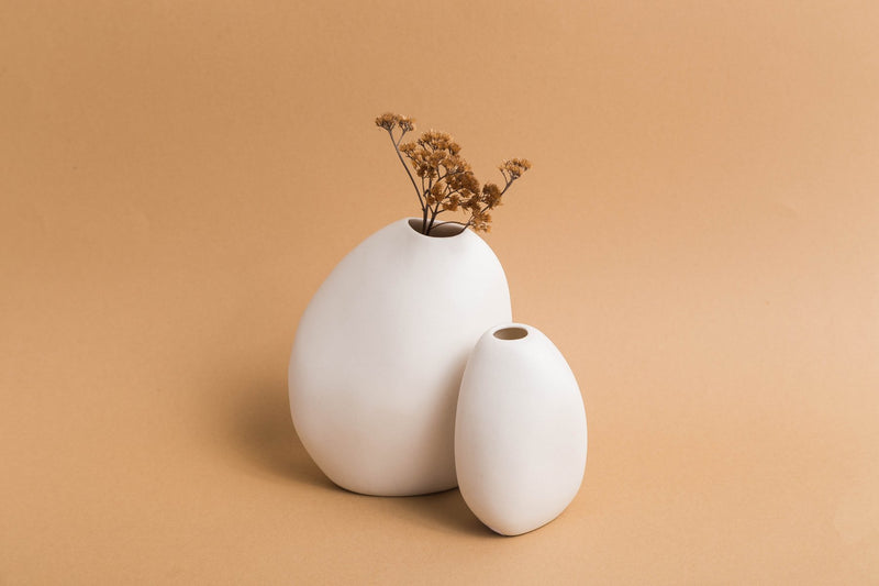 Two Harmie Vases, created by Vietnamese artisans, showcasing organic seed-like shapes, placed on a beige background. The vases are called Harmie Vase - Seed Grey and they are from the brand Ned Collections.