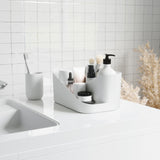 A white bathroom with a sink and various items on the counter. The counter is equipped with storage compartments and an Umbra Glam Cosmetic Organizer for makeup organization.