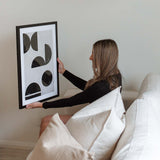 A woman sitting on a couch holding a black and white PAPIER HQ | GEOMETRIC PRINT by Art Prints, anxiously waiting for its delivery.