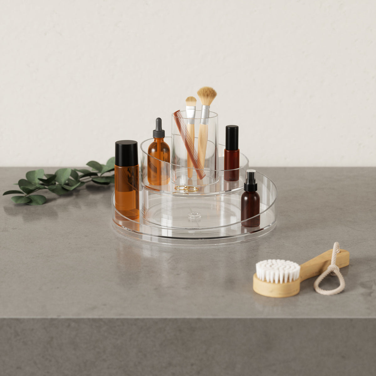 A clear tray with brushes and cosmetics on a table, featuring the Umbra Cascada Cosmetic Organizer with a rotating base.