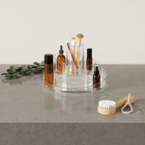 A clear tray with brushes and cosmetics on a table, featuring the Umbra Cascada Cosmetic Organizer with a rotating base.