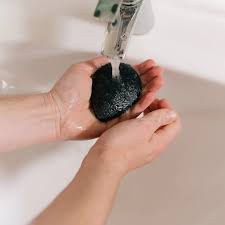 A person using a Florence Konjac Premium Facial Puff Sponge with Bamboo Charcoal to deep clean pores while washing it in a sink.