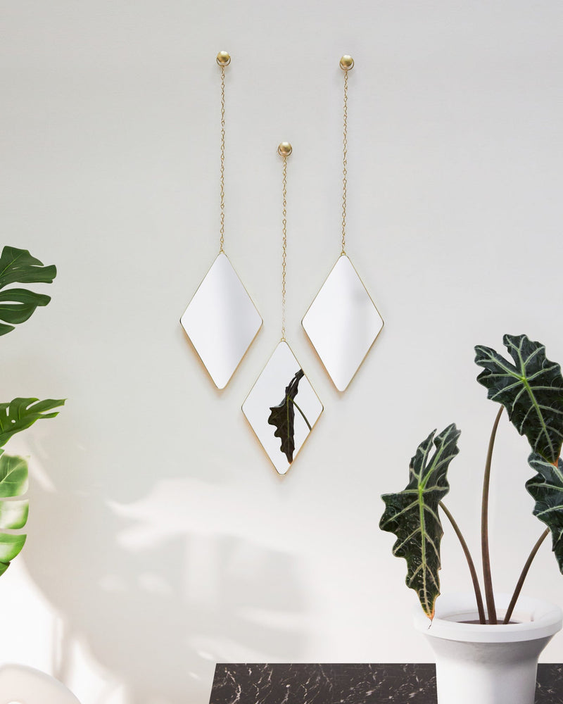 Three DIMA MIRROR - BRASS hanging mirrors with argyle patterns on a wall next to a potted plant. (Brand Name: Umbra)