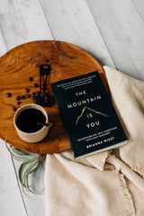 The Mountain Is You | Brianna Wiest" by Thought Catalog is on a wooden plate with a cup of coffee.