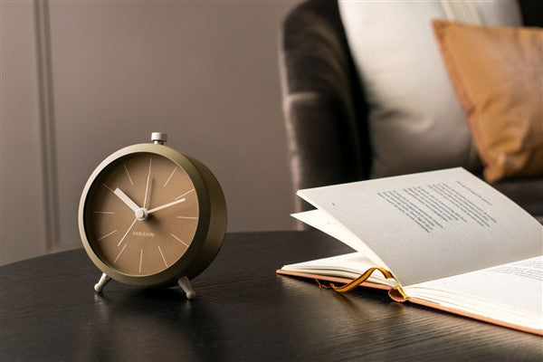 Moss green Button alarm clock on a dark bedside table beside an open book