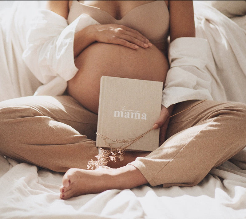 A pregnant woman sitting on a bed holding the Becoming MAMA - A Pregnancy Journal, the perfect diary gift for a baby shower.