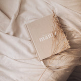 A Becoming MAMA - A Pregnancy Journal by AXEL & ASH showcasing a transformational journey and divine feminine strength, with the book titled "mama" placed on a bed.