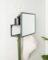 A Cubiko Mirror & Organizer - Black by Umbra, essential for small space living, hanging on a wall next to a plant and a hat.