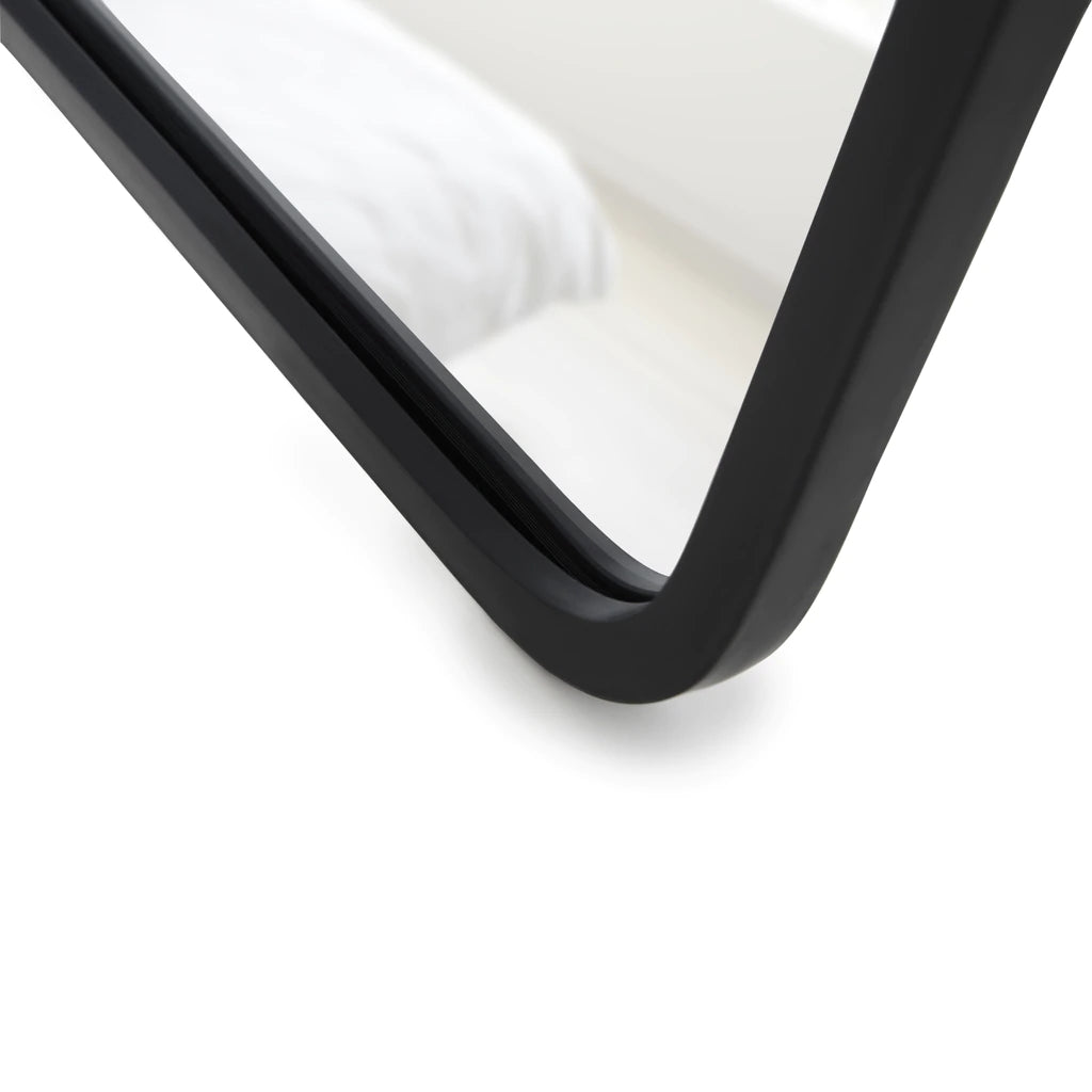 A close up of a full-length black HUB Leaning Mirror from the Umbra range on a white bed.