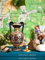 This table showcases a collection of memories and adventures, with a OUR TRAVELS. OUR FAMILY TRAVELS vase holding beautiful flowers, books filled with travel tales, and a map highlighting past explorations. (Brand: Write To Me)
