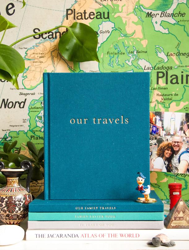Our Travels journal on a stack of books in front of a world map with photos and souvenirs