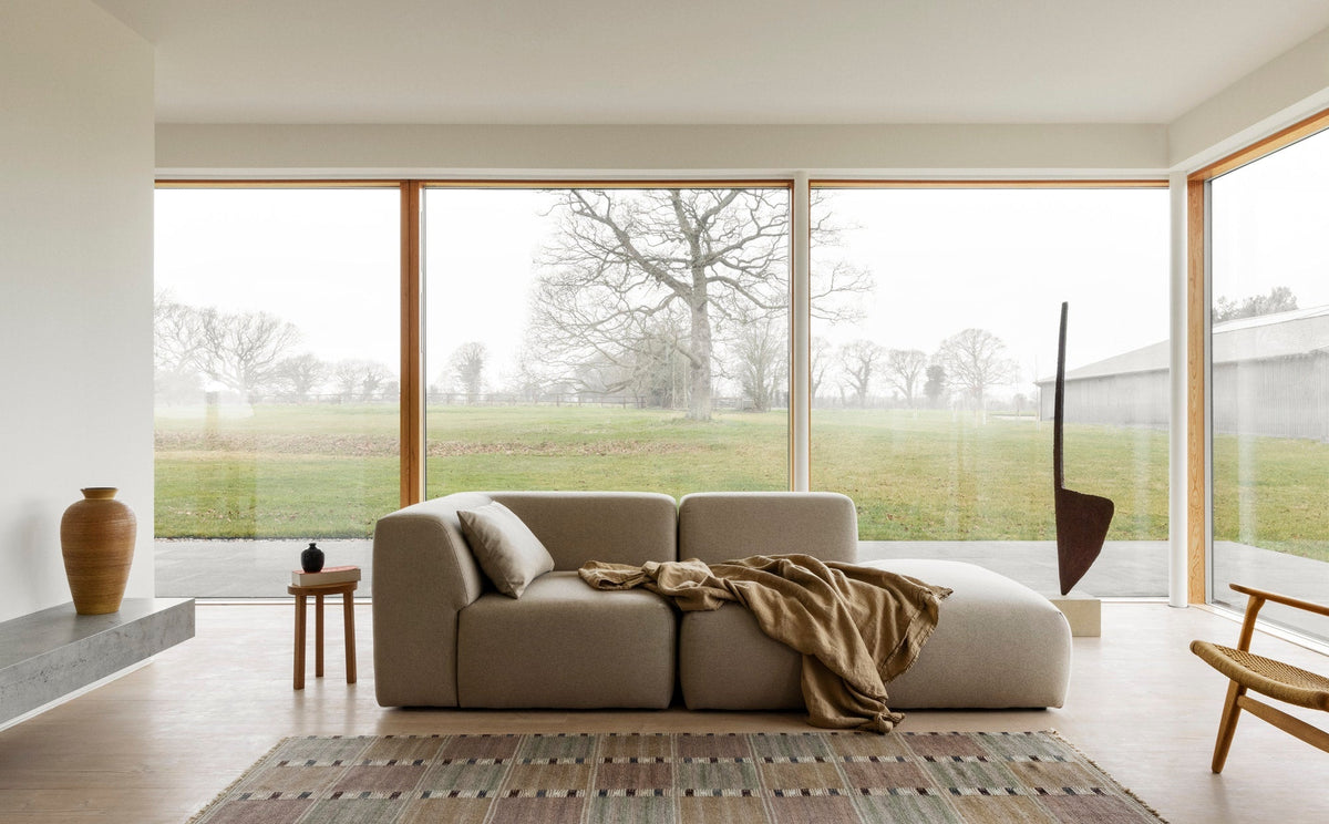 A minimal living room with a pale modular sofa, a woven rug and windows onto a field