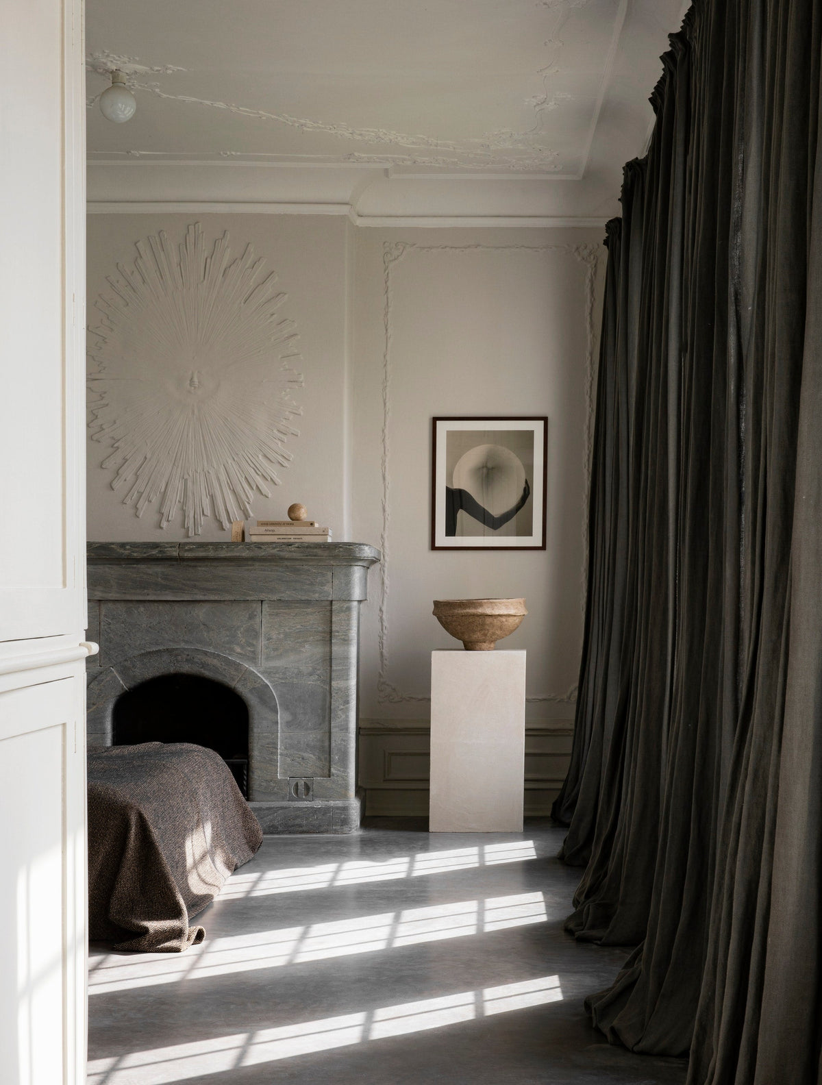 A pale classical room with a marble fireplace, plaster wall relief, dark curtains and sunlit floor