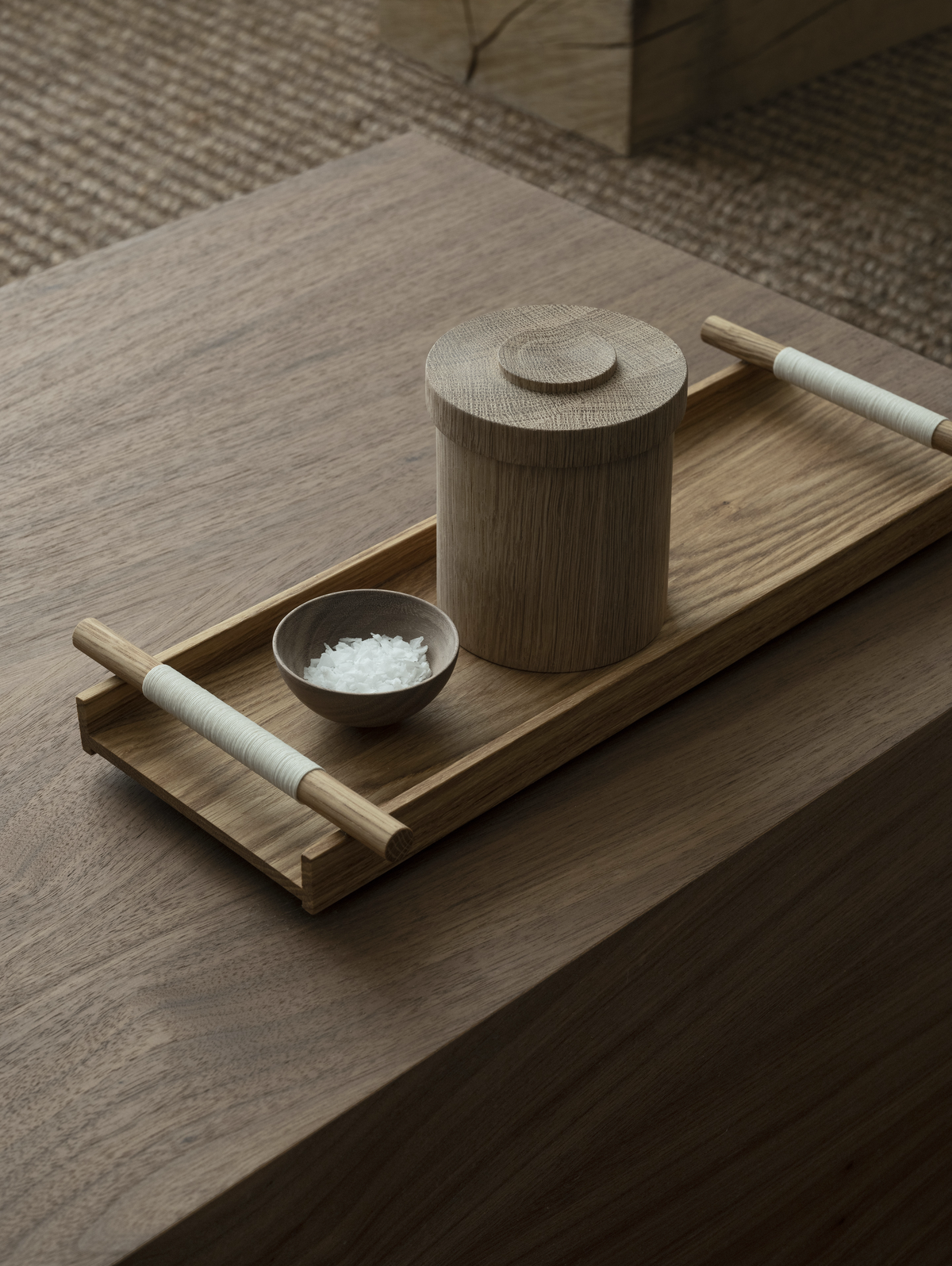 A wooden tray with turned handles holding a lidded canister and a small dark bowl on oak