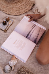 A woman is enjoying self-care on a blanket with the Healthy Nourished Soul - Book by Epicurean Publishing and seashells.