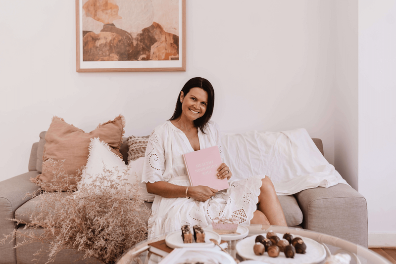 A woman in a white dress enjoying a cup of coffee on a couch as part of her self-care routine with the Healthy Nourished Soul - Book by Epicurean Publishing.