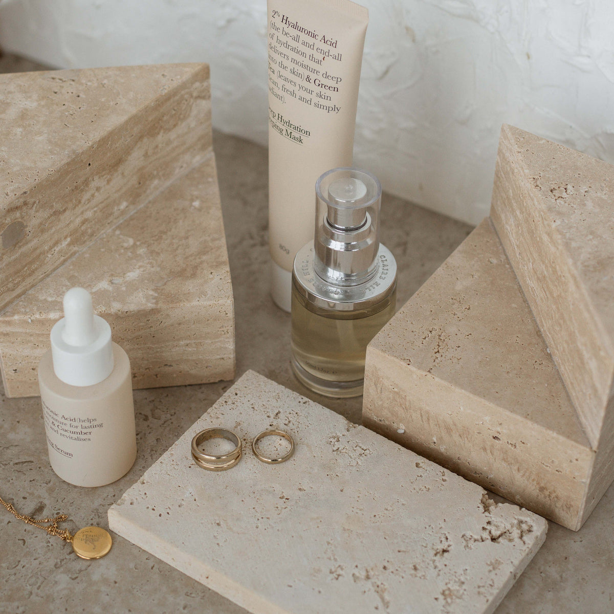 Two travertine wedge bookends styled with skincare bottles, gold rings and a travertine tray on a stone surface
