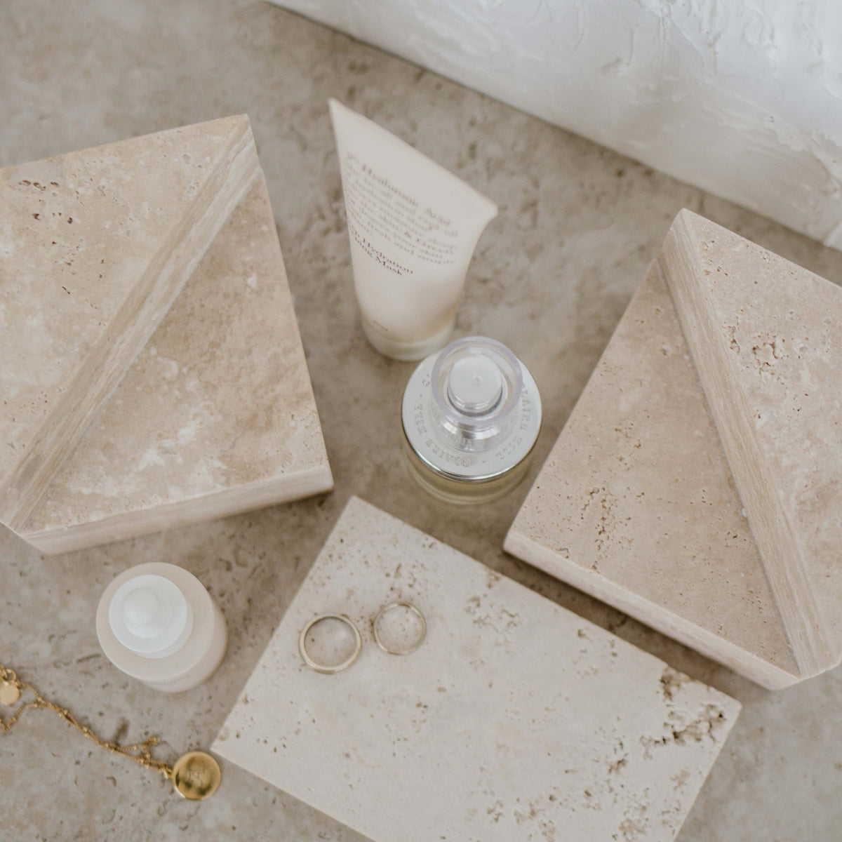 Travertine wedge bookends seen from above with skincare, a glass bottle, gold rings and a travertine tray