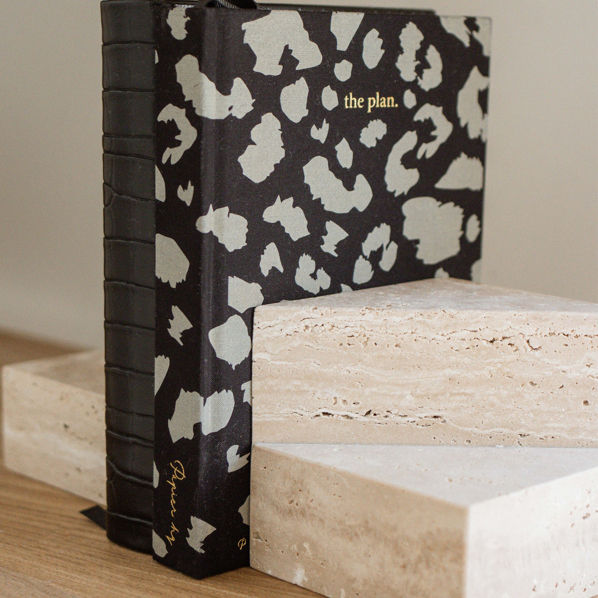 A travertine bookend holding up a patterned notebook titled the plan on a timber shelf