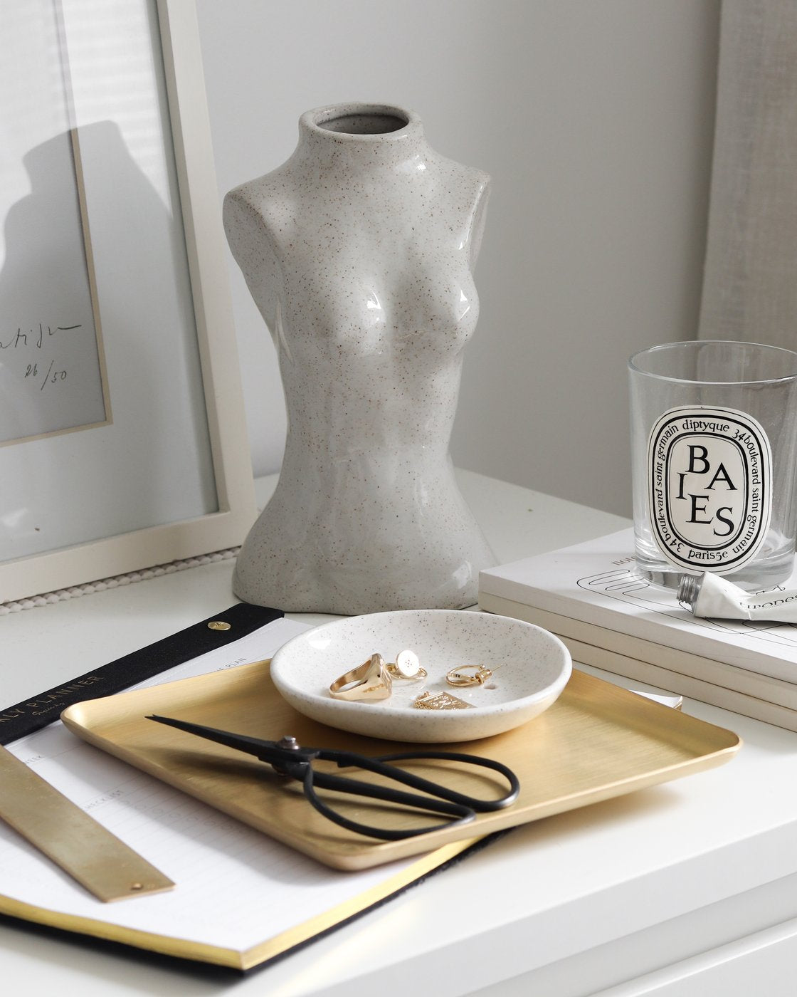 Speckled female silhouette vase on a white desk by a brass tray, jewellery dish, scissors and candle