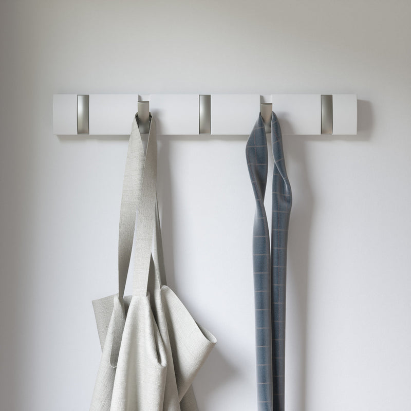 A wall mounted coat rack, the Umbra Flip 5 Hook White with retractable hooks for space-saving functionality, displaying a white coat hanger with a grey apron hanging on it.