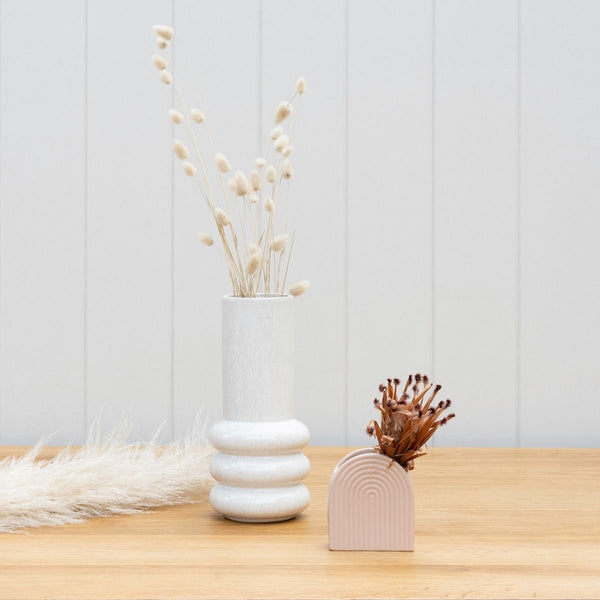 The Potted Milan Vase, a stunning décor statement, showcases a white vase with a pink flower gracefully resting on top of a wooden table.