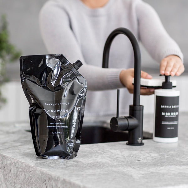 A woman pouring Barkly Basics Dish Wash Refill into a black bag on a kitchen counter.