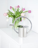 A Quench Watering Can - Stainless Steel, from the Umbra range, sits on a table next to a vase of tulips. It features a 360-degree handle for easy use.